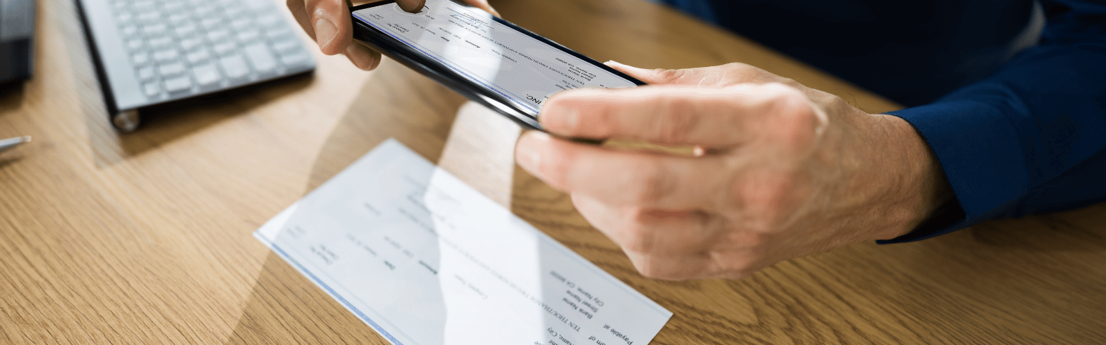 Person holding a tablet over a wooden desk, with a printed check and papers nearby and a keyboard in the background.